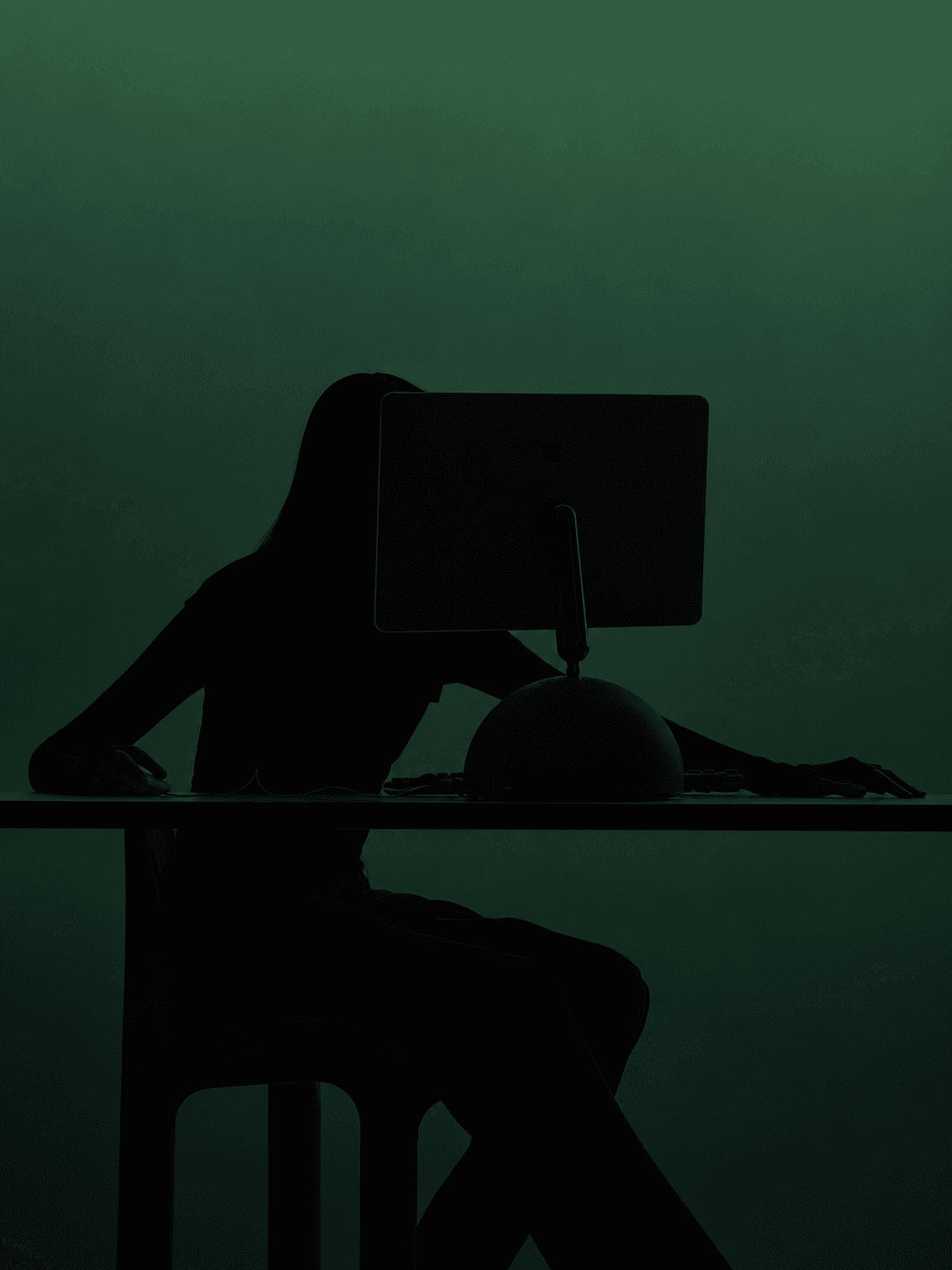Silhouette of a person seated at a desk, working behind a desktop computer against a green backdrop.